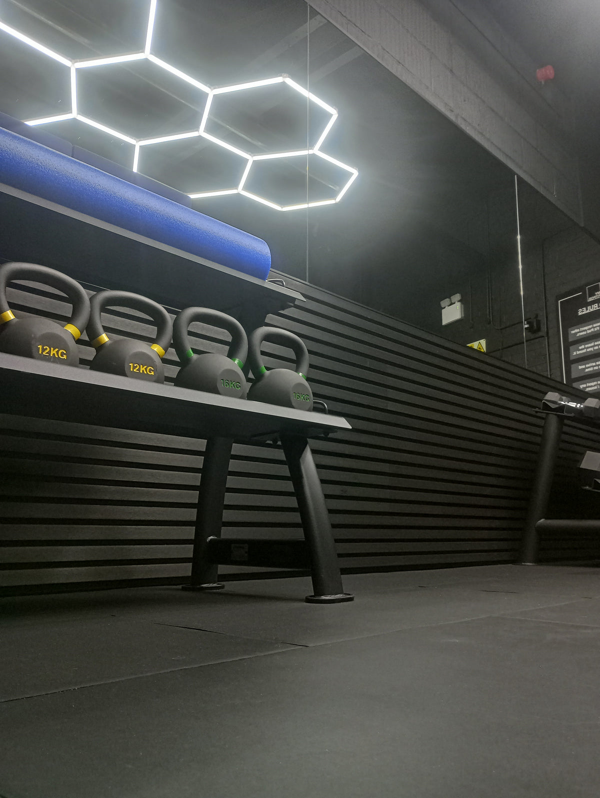 Four black kettlebells with yellow and green markings resting on a black weight bench in a modern gym setting with a dark slatted wall and hexagonal ceiling lights.